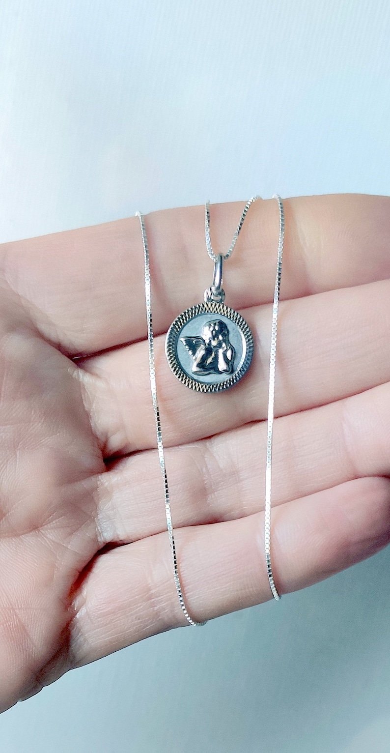 May include: A silver necklace featuring a round pendant with an angel design. The pendant has a detailed image of an angel within a textured border. The chain is a delicate, square-linked style. The necklace is displayed against a light background.