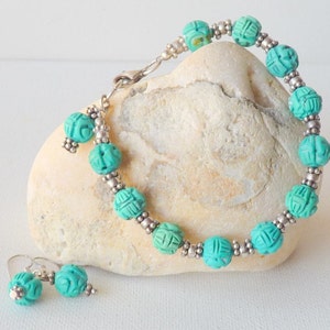 May include: A turquoise beaded bracelet and matching earrings set. The bracelet features carved turquoise beads separated by small silver-colored beads. The earrings have a similar design. The jewelry is displayed on a light-colored stone.