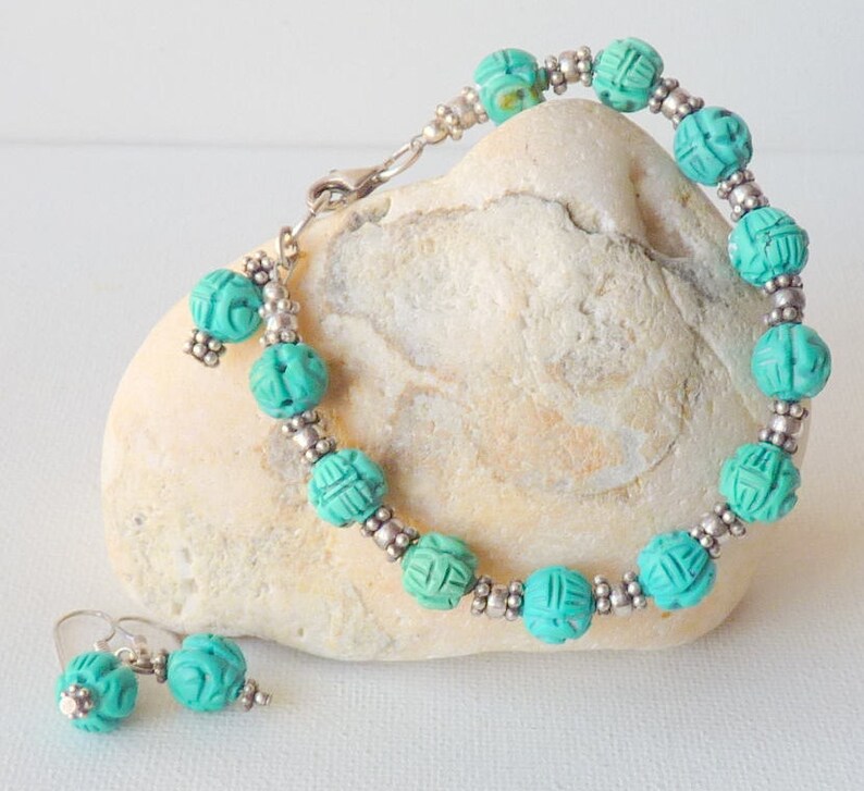 May include: A turquoise beaded bracelet and matching earrings set. The bracelet features carved turquoise beads separated by small silver-colored beads. The earrings have a similar design. The jewelry is displayed on a light-colored stone.