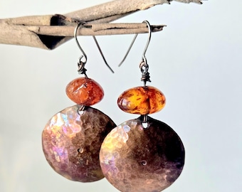 May include: A pair of drop earrings featuring a round, hammered copper disc suspended below a smooth, translucent amber-colored bead. The earrings are attached to silver-toned metal ear wires, showcasing a contrast between the metallic texture and the warm, polished stone elements.