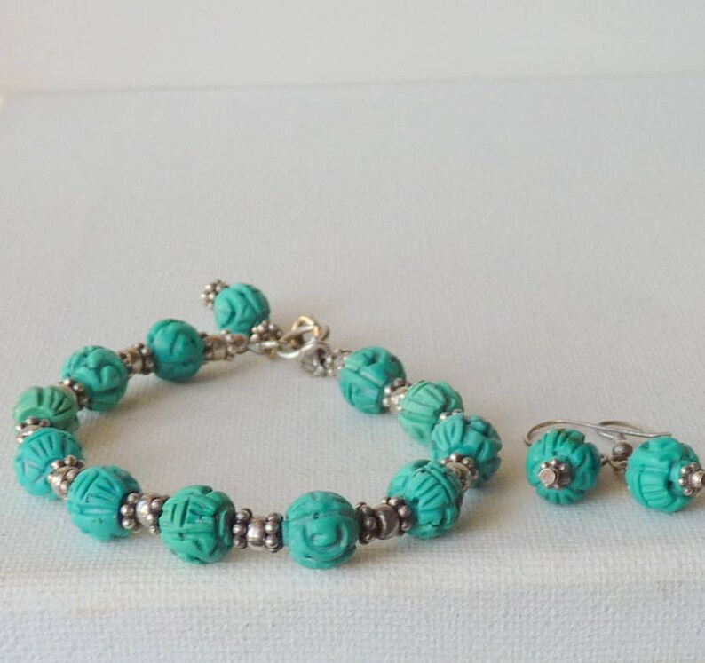 May include: A turquoise beaded bracelet and matching earrings set. The bracelet features carved round beads with silver-toned spacers. The earrings have similar carved turquoise beads. The jewelry set is displayed on a white surface.