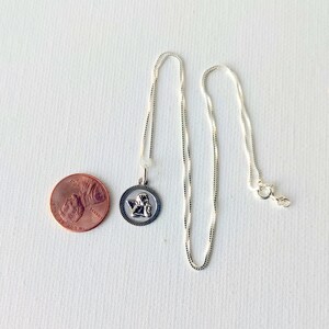 May include: A silver-colored necklace with a round pendant featuring an angel design. The pendant is next to a penny for size comparison. The necklace chain is delicate and has a lobster clasp.