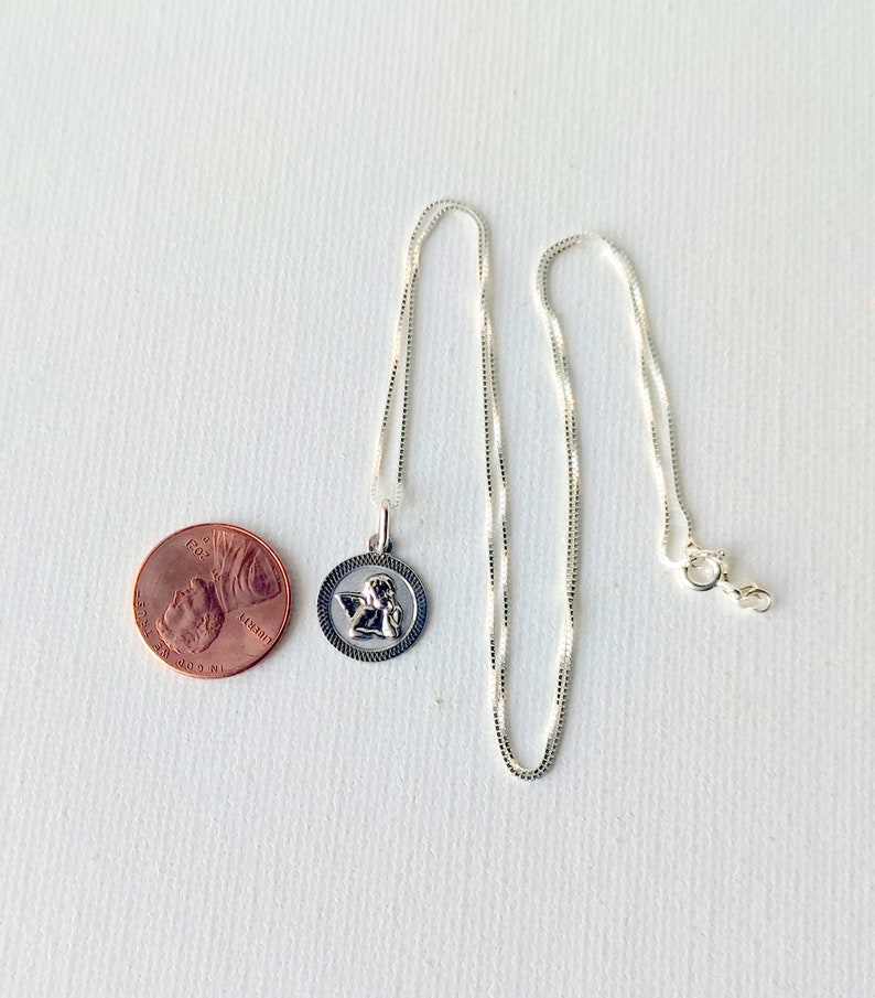 May include: A silver-colored necklace with a round pendant featuring an angel design. The pendant is next to a penny for size comparison. The necklace chain is delicate and has a lobster clasp.