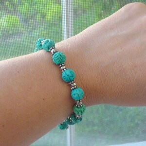 May include: A turquoise beaded bracelet with carved spherical beads and silver-colored spacers. The bracelet is worn on a wrist, showcasing the intricate details of the carved design and the contrast between the turquoise and silver elements.