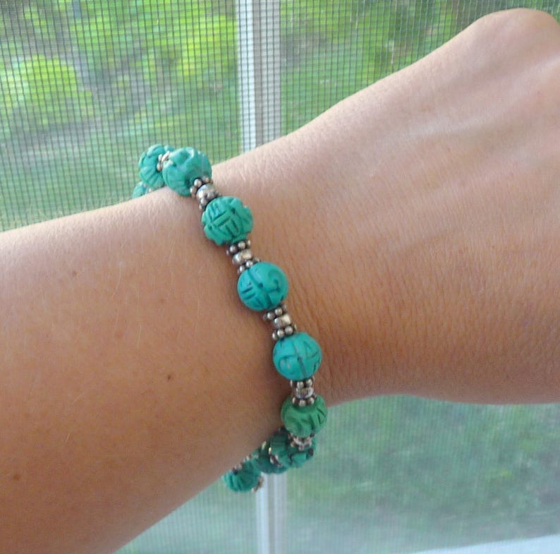 May include: A turquoise beaded bracelet with carved spherical beads and silver-colored spacers. The bracelet is worn on a wrist, showcasing the intricate details of the carved design and the contrast between the turquoise and silver elements.