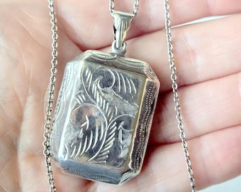 May include: A rectangular silver locket pendant with beveled edges and intricate engraved floral patterns on the front surface. The locket is attached to a delicate silver chain link necklace that extends vertically across the frame.