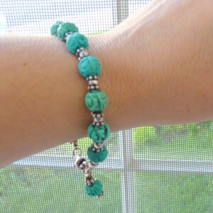 May include: A turquoise beaded bracelet with carved floral designs. The bracelet features round beads separated by silver-colored spacers. The bracelet is secured with a silver-colored clasp and is displayed on a person's arm.