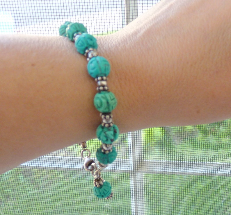 May include: A turquoise beaded bracelet with carved floral designs. The bracelet features round beads separated by silver-colored spacers. The bracelet is secured with a silver-colored clasp and is displayed on a person's arm.