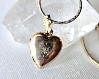 May include: A gold-toned heart-shaped locket pendant suspended from a silver-toned snake chain. The locket features a small, centered starburst engraving with a tiny embedded clear stone. The pendant has a decorative bail at the top.