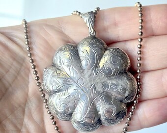 May include: A silver-toned metal locket pendant shaped like a six-petaled flower with intricate engraved scrollwork and floral patterns. The pendant is suspended from a matching silver-toned ball chain necklace with a textured metal bail.