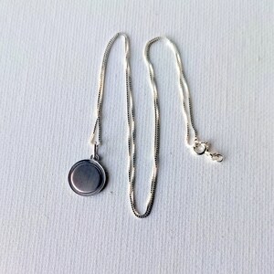 May include: A silver-colored necklace with a round pendant. The pendant is a flat disc shape. The chain is a delicate box chain style, with a lobster clasp closure. The necklace is displayed on a white surface.