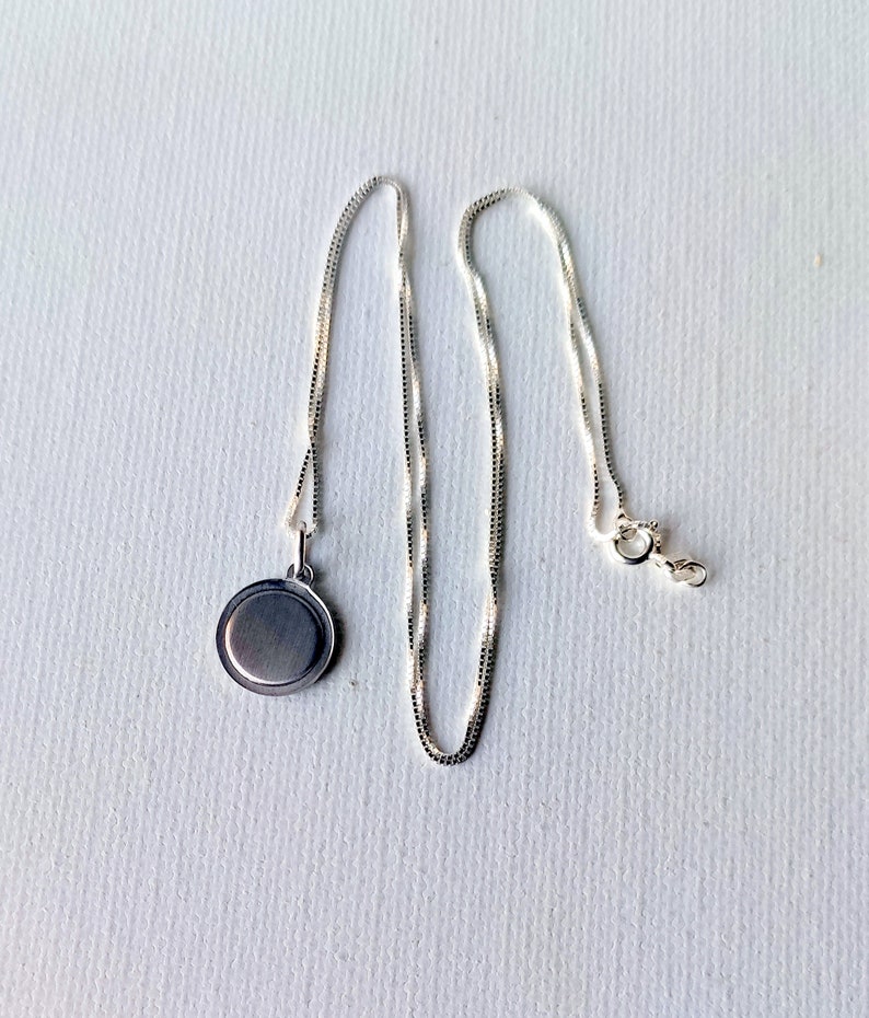 May include: A silver-colored necklace with a round pendant. The pendant is a flat disc shape. The chain is a delicate box chain style, with a lobster clasp closure. The necklace is displayed on a white surface.