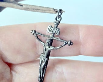 May include: A silver-toned metal crucifix pendant featuring a detailed figure of Jesus on the cross. The top of the cross displays the letters INRI on a small plaque. The pendant includes a small circular jump ring at the top for attachment.