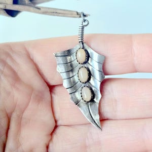 May include: A silver-toned metal pendant shaped like a leaf with a textured, ribbed surface. The pendant features three vertically aligned, oval-shaped, light-colored cabochon stones set in serrated bezels along the center vein of the leaf.