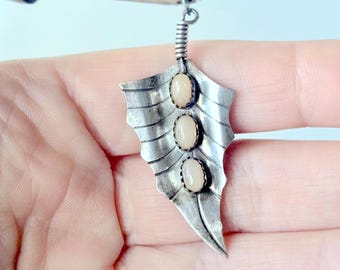 May include: A silver-toned metal pendant shaped like a leaf with a textured, ribbed surface. The pendant features three vertically aligned, oval-shaped, light-colored cabochon stones set in serrated bezels along the center vein of the leaf.