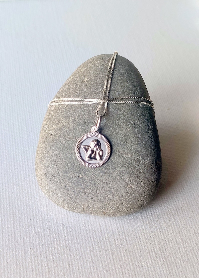 May include: A silver-colored pendant necklace featuring a round charm with a detailed angel design. The charm is encircled by a decorative border. The necklace is displayed on a gray stone against a white background.