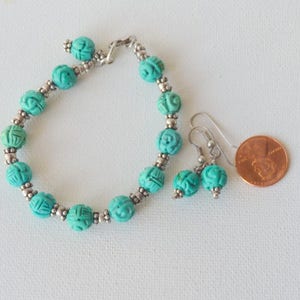 May include: A turquoise beaded bracelet and matching earrings set. The bracelet and earrings feature carved round beads with silver-colored accents. A penny is next to the jewelry for size comparison. The jewelry is on a white surface.