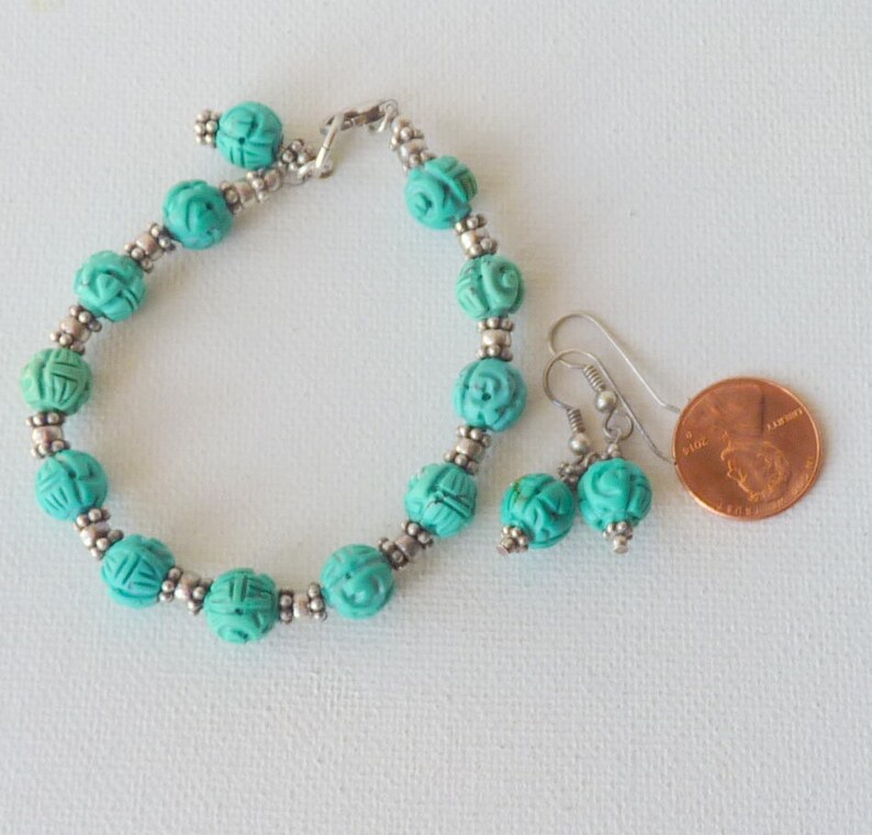 May include: A turquoise beaded bracelet and matching earrings set. The bracelet and earrings feature carved round beads with silver-colored accents. A penny is next to the jewelry for size comparison. The jewelry is on a white surface.