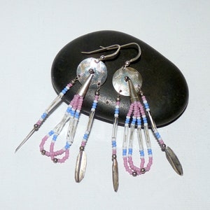 May include: Silver dangle earrings with a circular, textured top and beaded strands in pink, light blue, and clear. The earrings have silver cone-shaped accents and feather-shaped dangles. The earrings are displayed on a dark gray stone.