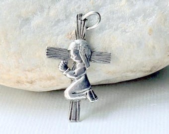 May include: A silver-toned metal pendant shaped like a cross, featuring a sculpted figure of a person kneeling in prayer. The cross has a textured, linear design on its arms and vertical post, with a small circular bail at the top for hanging.