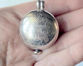 May include: A round, silver-colored metal pendant featuring an intricate, engraved design of a stylized basket or vessel surrounded by cloud-like patterns. The circular object has a small loop at the top and a rounded decorative knob at the bottom.