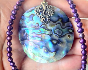 May include: A necklace featuring a large, round, iridescent abalone shell pendant with a silver-toned metal filigree bail, strung on a strand of small, round, deep purple pearls with a silver-toned metal clasp closure.