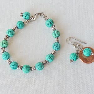 May include: A turquoise beaded bracelet and matching earrings set. The bracelet and earrings feature carved beads with silver-colored accents. The earrings have a hook closure. The jewelry is displayed on a white surface.