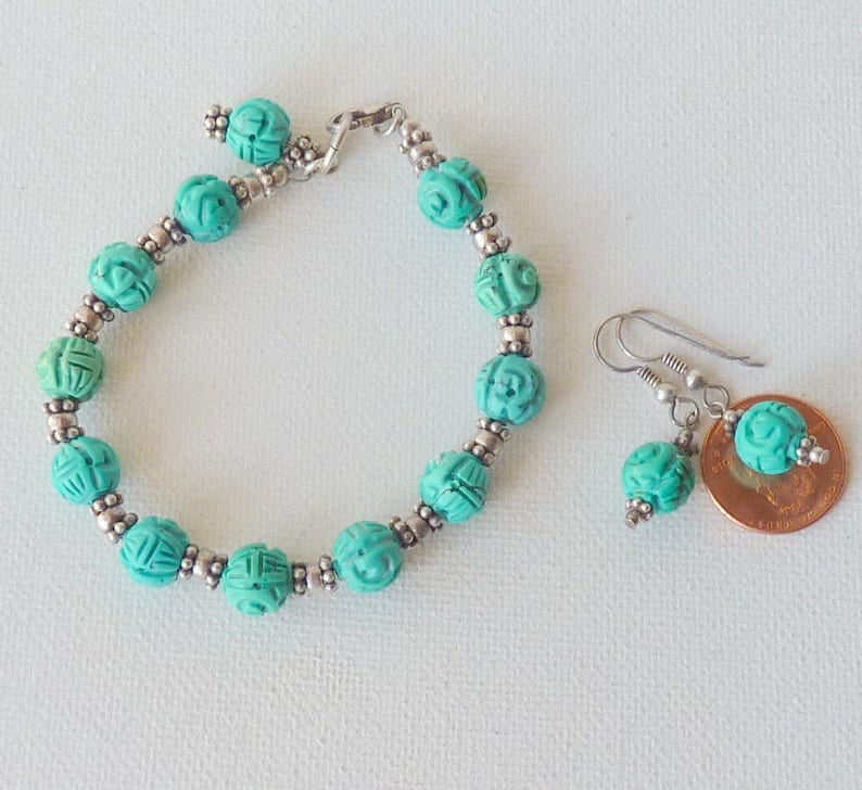 May include: A turquoise beaded bracelet and matching earrings set. The bracelet and earrings feature carved beads with silver-colored accents. The earrings have a hook closure. The jewelry is displayed on a white surface.