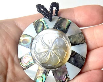 May include: A circular pendant featuring a central carved mother-of-pearl disc with a floral motif, surrounded by alternating segments of white shell and iridescent abalone shell. The pendant is attached to a black beaded cord at the top.