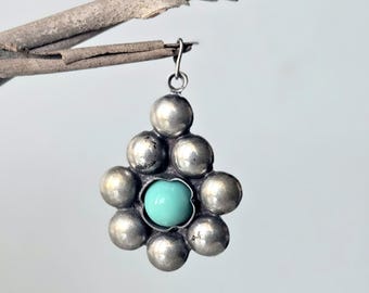 May include: A silver pendant featuring a cluster of eight polished spherical silver beads arranged in a flower-like pattern around a single, smooth, round, light turquoise-colored stone set in the center, complete with a small silver jump ring.