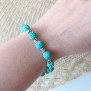 May include: A turquoise beaded bracelet with carved details and silver-colored spacer beads. The bracelet is worn on a wrist, showcasing the intricate design and color. The beads are approximately 0.5 inches in diameter, creating a striking accessory.