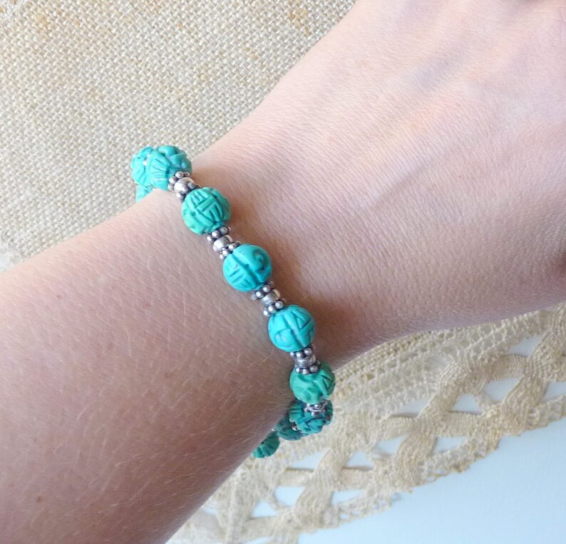 May include: A turquoise beaded bracelet with carved details and silver-colored spacer beads. The bracelet is worn on a wrist, showcasing the intricate design and color. The beads are approximately 0.5 inches in diameter, creating a striking accessory.
