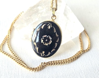 May include: An oval-shaped gold locket pendant featuring a smooth black inlay with a central flower motif and surrounding leaf-like engravings. The locket is suspended from a delicate gold chain, showcasing a polished finish and classic design.