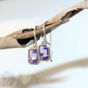 May include: A pair of silver earrings with rectangular, light purple gemstones. The earrings are suspended from simple hooks and are displayed on a small, light brown branch against a soft, blurred background.