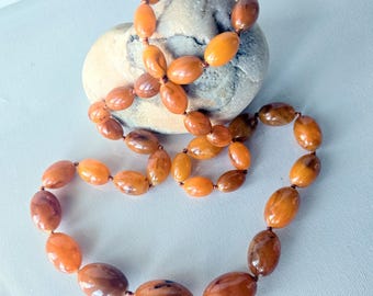 May include: A necklace composed of smooth, oval-shaped beads in varying shades of translucent orange, amber, and brown. The beads are strung together on a thin cord, featuring a polished, glossy finish that reflects light across their rounded surfaces.