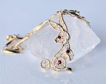 May include: A gold-toned metal necklace featuring a rabbit-shaped pendant with intricate filigree cutouts. The rabbit design is accented with three small, round, dark red gemstones embedded in the head, body, and tail areas of the pendant.