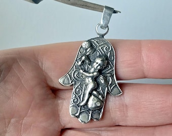 May include: A silver-toned Hamsa pendant featuring an embossed cherub figure in the center. The hand-shaped charm has intricate swirling patterns in the background and a small metal loop at the top for attachment to a necklace or chain.