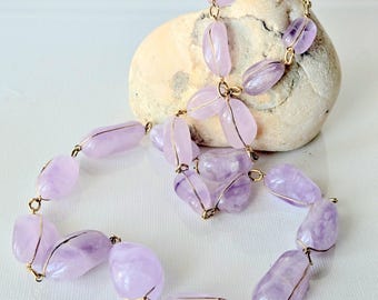 May include: A necklace featuring smooth, polished, light purple amethyst stones, each individually wrapped in thin gold-toned wire. The stones are connected by small gold-toned metal jump rings, creating a continuous chain of rounded, translucent purple gems.