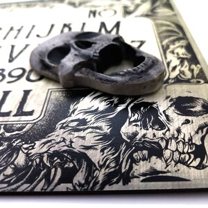May include: A wooden Ouija board with black and white illustrations of skulls and wolves. The board has the letters of the alphabet, numbers 1-9 and 0, and the words "yes", "no", "goodbye", and "hello". A gray skull-shaped planchette is on top of the board.