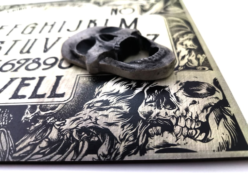 May include: A wooden Ouija board with black and white illustrations of skulls and wolves. The board has the letters of the alphabet, numbers 1-9 and 0, and the words "yes", "no", "goodbye", and "hello". A gray skull-shaped planchette is on top of the board.