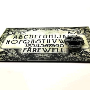 May include: A black and white Ouija board with a skull planchette. The board has the letters of the alphabet, numbers 1-9 and 0, and the words "YES", "NO", and "FAREWELL". The board is surrounded by a border of skulls.