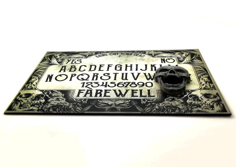 May include: A black and white Ouija board with a skull planchette. The board has the letters of the alphabet, numbers 1-9 and 0, and the words "YES", "NO", and "FAREWELL". The board is surrounded by a border of skulls.