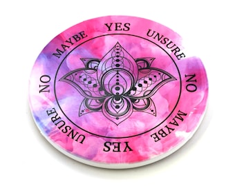 Op de afbeelding: Een roze en paarse cirkelvormig divinatiebord met een zwart lotusmandala-ontwerp. Het bord heeft de woorden "JA", "MISSCHIEN", "NEE", "ONZEKER" rond de rand.