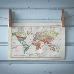 Op de afbeelding: Een vintage wereldkaart gedrukt op papier met een lichtblauwe achtergrond. De kaart toont de continenten in verschillende kleuren, waaronder rood, oranje, geel, groen en blauw. De kaart heeft de titel "Statistisch Atlas en Gids".