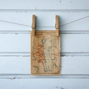 May include: A vintage map of St. Augustine, Florida, is held by two wooden clothes pegs on a wire against a white, weathered wood background. The map is tan with red and black details.