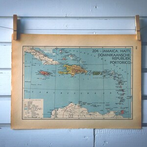 May include: A vintage map print of Jamaica, Haiti, Dominican Republic, and Puerto Rico, displayed against a light blue wooden backdrop. The map is held by two wooden pegs. The map features a detailed illustration with various colours.