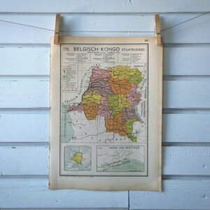Puede incluir: Un mapa antiguo del Congo Belga, titulado "176. Belgisch Kongo Staatkundig", está suspendido por pinzas de madera contra una pared de madera azul claro. El mapa presenta una ilustración detallada y colorida de la región.