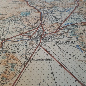May include: Close-up of a vintage map with detailed cartography. The map features a network of roads, rivers, and topographical details, with the city of Podgorica prominently labeled. The map's color palette includes beige, brown, and red, with text in a foreign language.