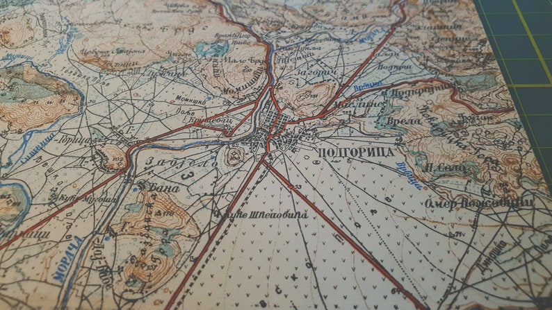 May include: Close-up of a vintage map with detailed cartography. The map features a network of roads, rivers, and topographical details, with the city of Podgorica prominently labeled. The map's color palette includes beige, brown, and red, with text in a foreign language.