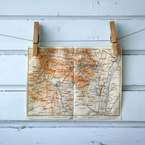 Puede incluir: Un mapa antiguo, con tonos marrones y beige, está suspendido por dos pinzas de madera sobre un fondo de madera azul claro desgastado. El mapa muestra detalles geográficos y texto.